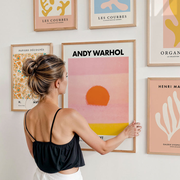 Woman arranging framed posters on a gallery wall with 'Andy Warhol' and other abstract art pieces by famous artists.