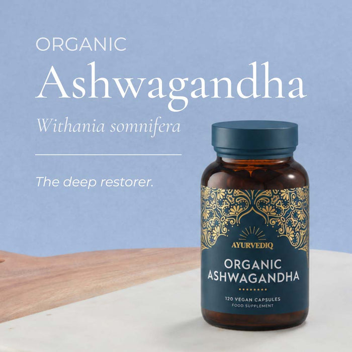 A bottle of Ayurvedic Organic Ashwagandha capsules with a dark blue label and gold ornamental pattern, set on a white surface with a wooden board. The background is a soft light blue with white text reading 'ORGANIC Ashwagandha Withania somnifera The deep restorer.