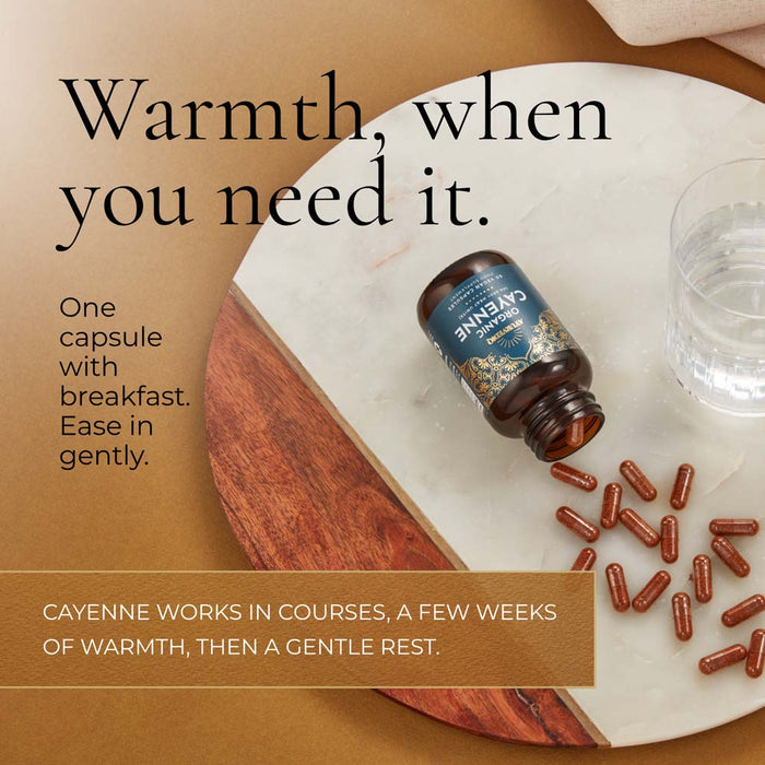 A bottle of Cayenne capsules lies on a white marble tray with several capsules scattered beside it, alongside a glass of water. Large text reads, 'Warmth, when you need it.'