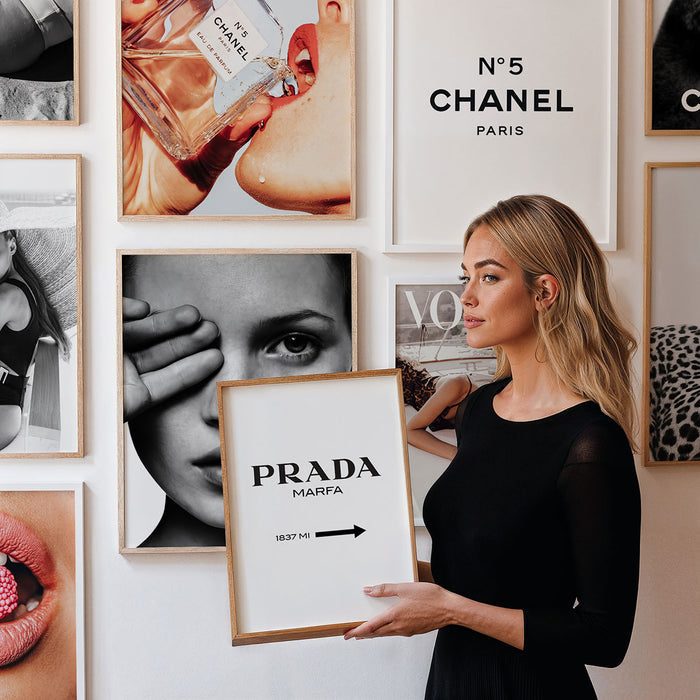 Woman holding a framed picture with 'Prada' text against a gallery wall of framed art and fashion images.