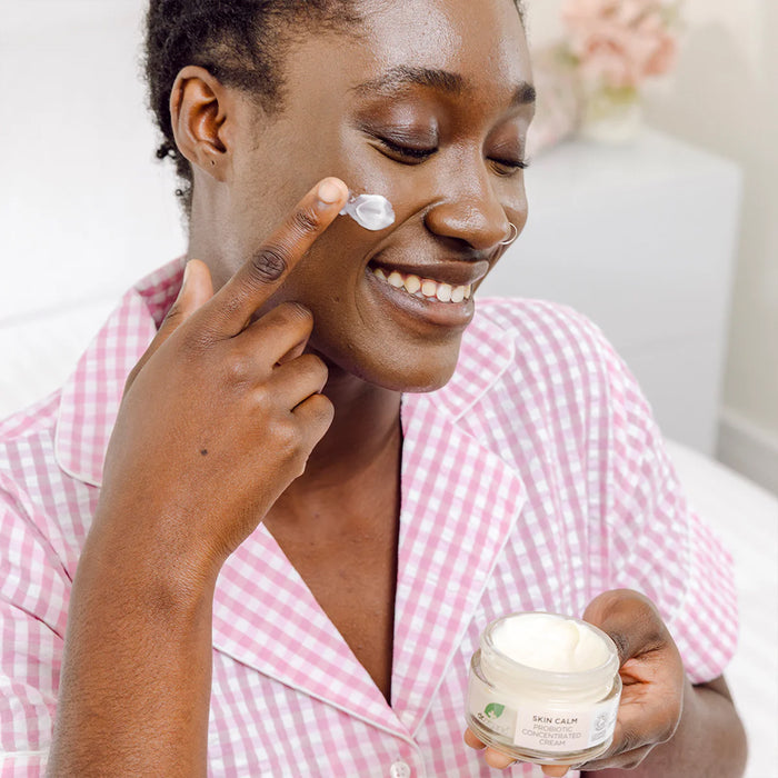 Woman applying Dr. Organic Skin Calm Probiotic Concentrated Cream to her face with a jar of cream in her hand, wearing a pink checkered shirt.