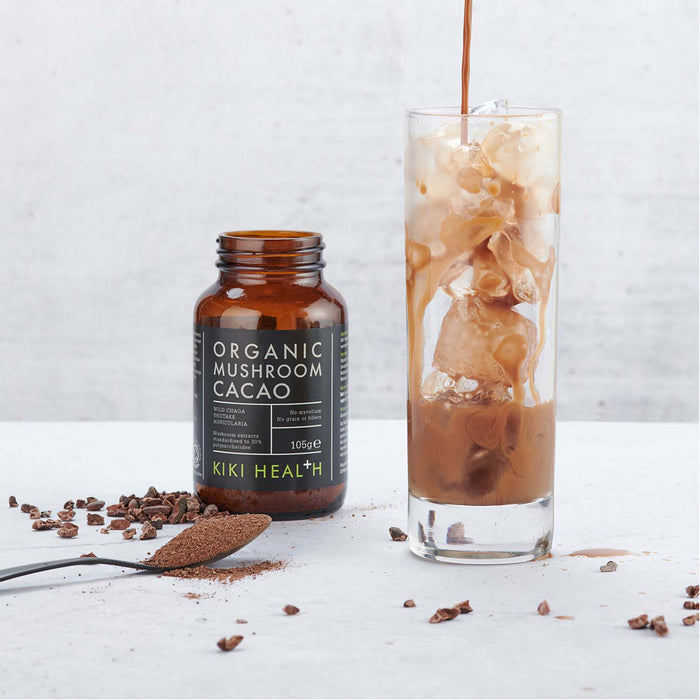 Kiki Health Organic Mushroom Cacao bottle with a glass of iced coffee and cocoa powder on a white surface.