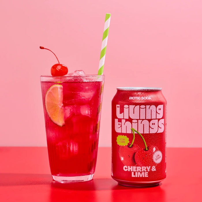 Red drink in a glass with a cherry and lime, and a can of Living Things Cherry & Lime soda on a pink background.