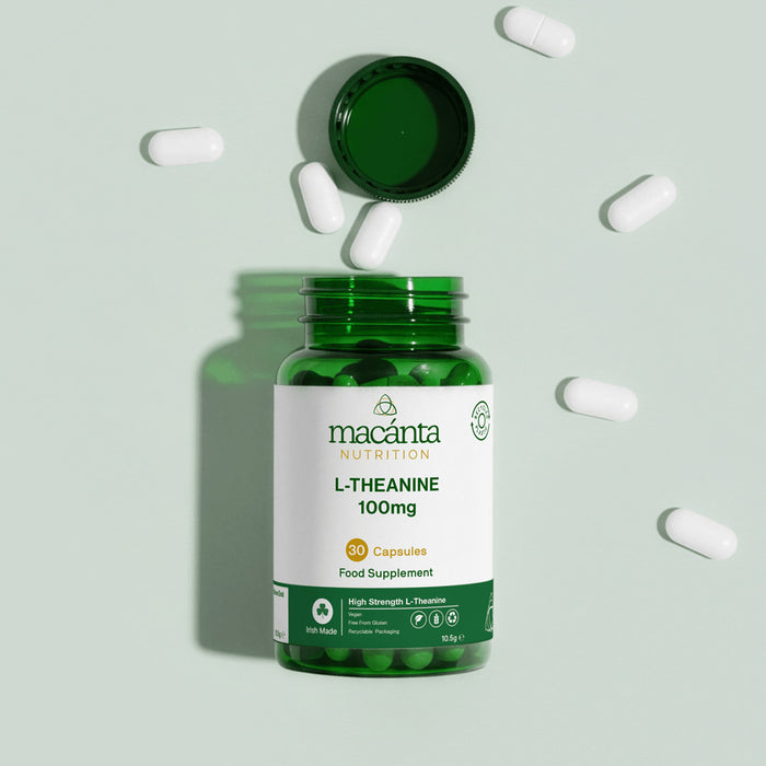 A green bottle of Macanta Nutrition L-Theanine 100mg capsules with the cap off, surrounded by scattered white capsules on a pale green background.