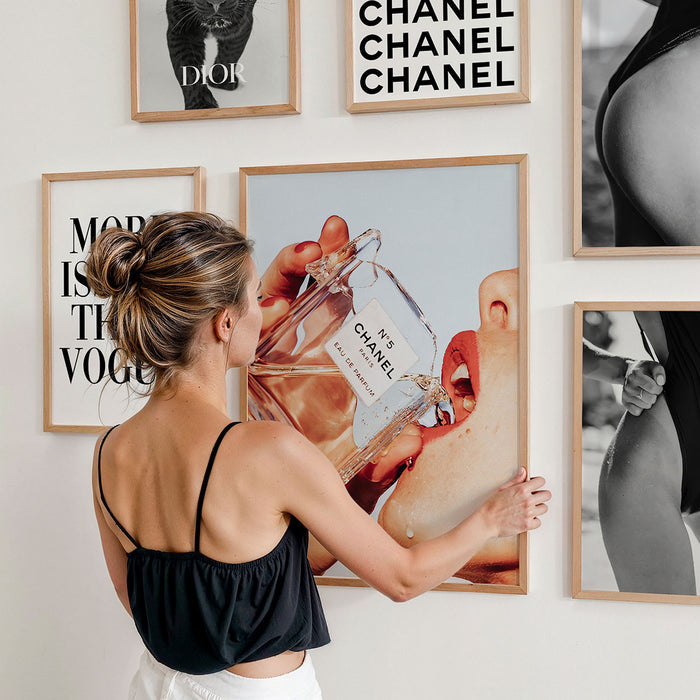 Woman adjusting a framed art print of a Chanel perfume bottle on a wall with other fashion themed wall art.