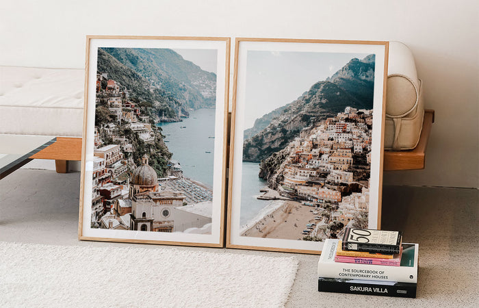 Two framed prints of a European coastal town on a white wall with books below.