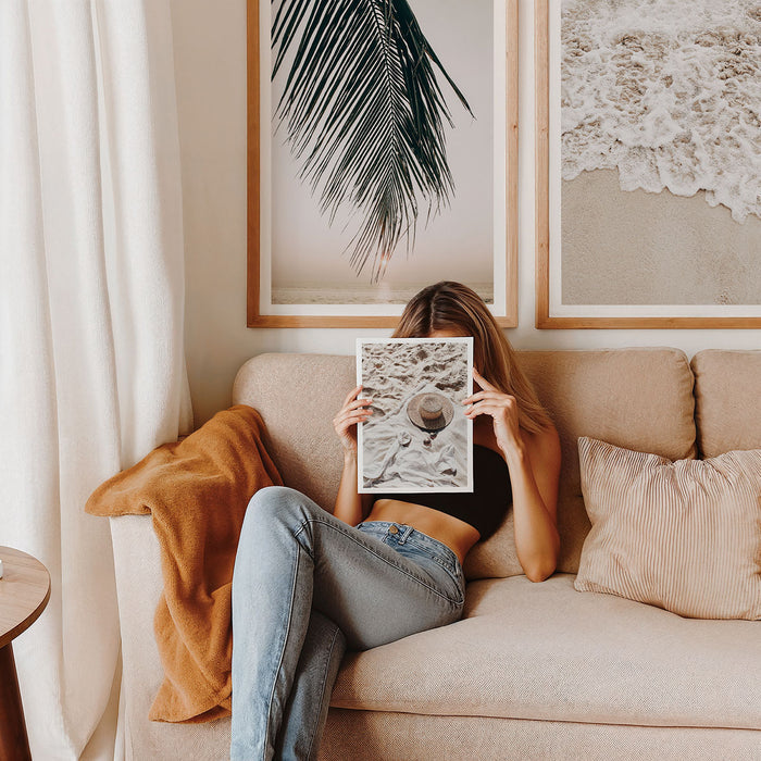 Person sitting on a couch holding an art print in front of their face, with two large framed prints in the background.