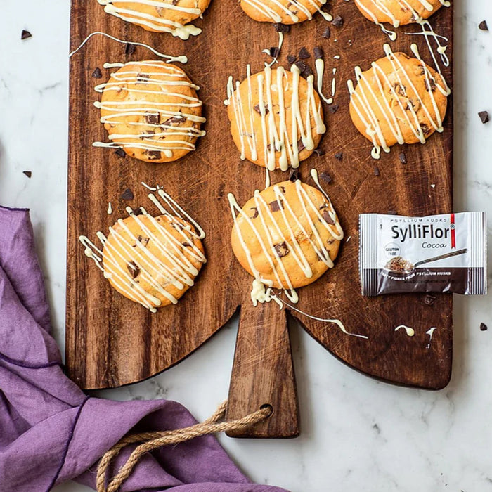 Chocolate chip cookies topped with white drizzle arranged on a wooden serving board, with a package of SylliFlor cocoa cookies and a purple cloth on a light marble surface.