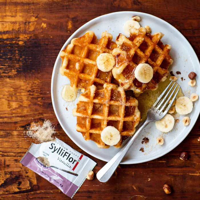 Three golden-brown waffles topped with banana slices and powdered sugar on a white plate, set on a rustic wooden table with a fork, hazelnuts, and a SylliFlor vanilla packet nearby.