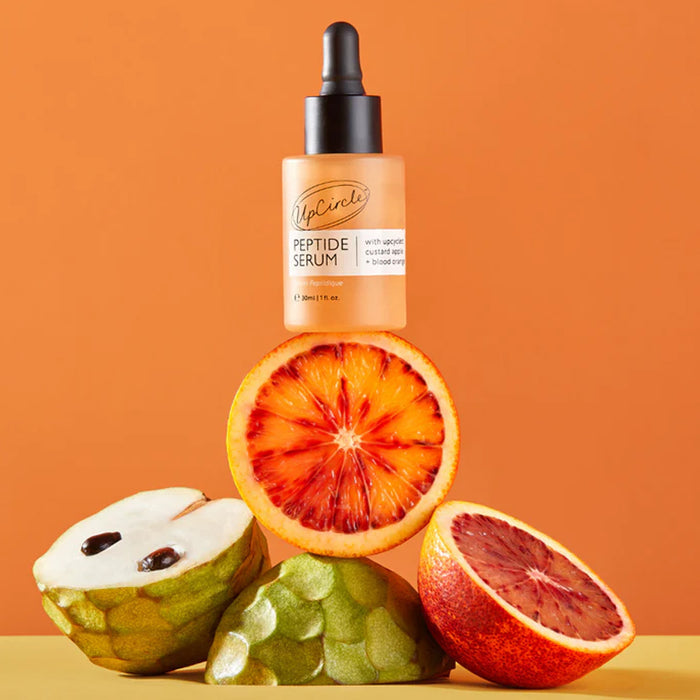 Peptide serum bottle with blood oranges and a green fruit on an orange background