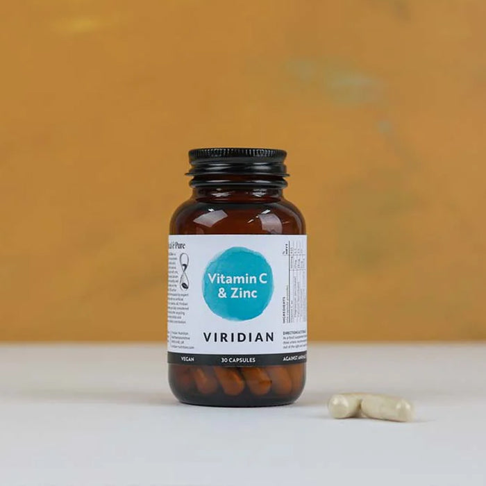 Brown supplement bottle labeled 'Vitamin C & Zinc' by Viridian on a white surface with a blurred orange background.