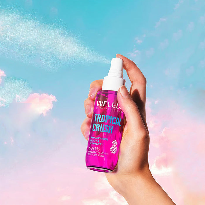 A hand holding a bottle of Weleda Naturals Tropical Crush Perfume Mist against a pastel sky background with pink clouds.