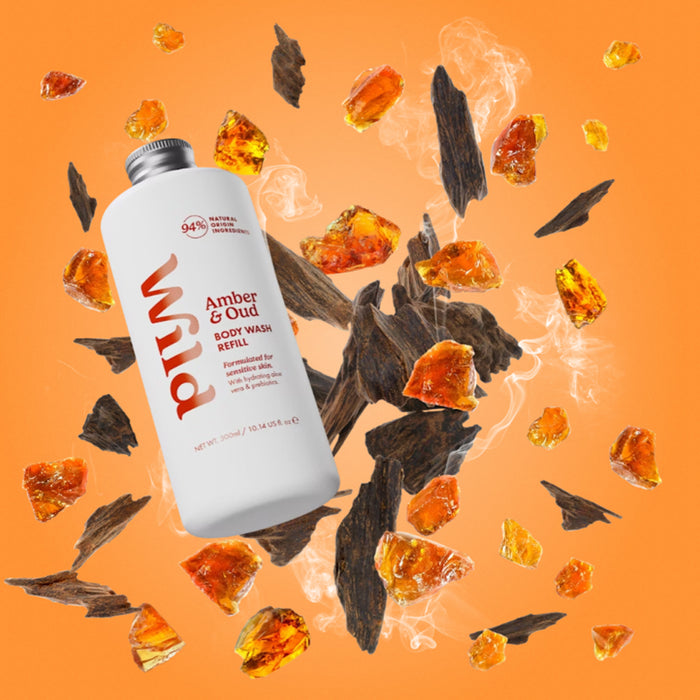 A bottle of Wild Amber & Oud Scrub Wash floating over scattered amber pieces and dark wood shavings on an orange background.